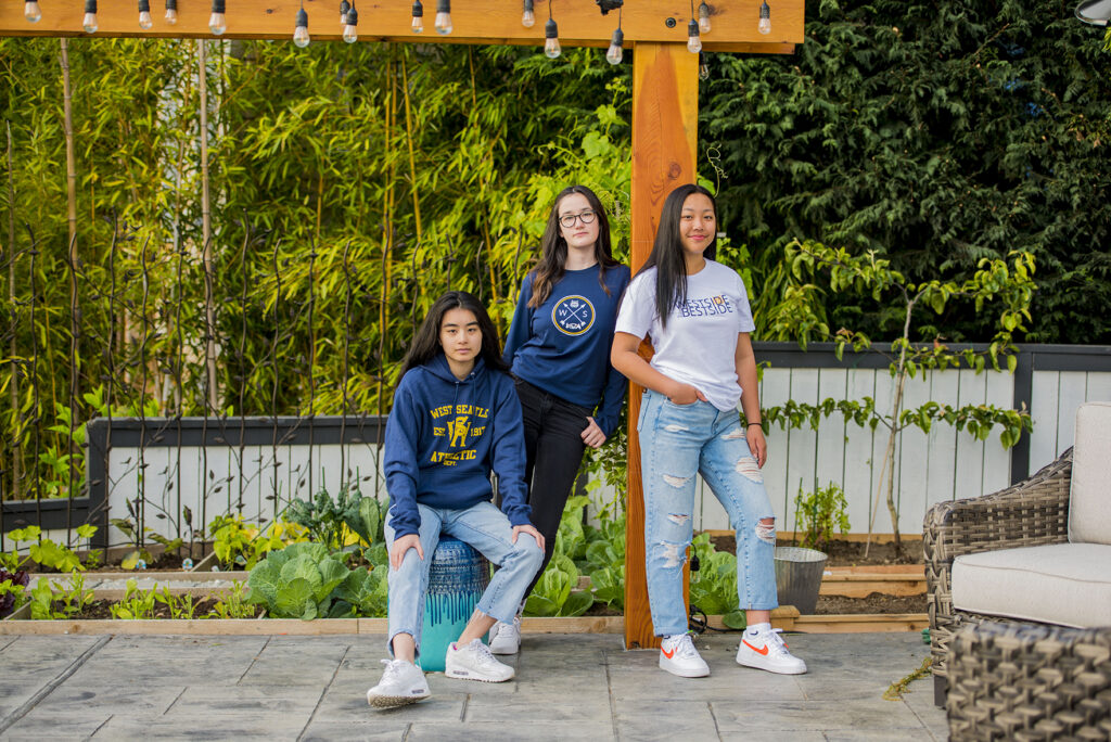 Three female student-athletes wearing West Seattle Athletics gear pose in a garden.